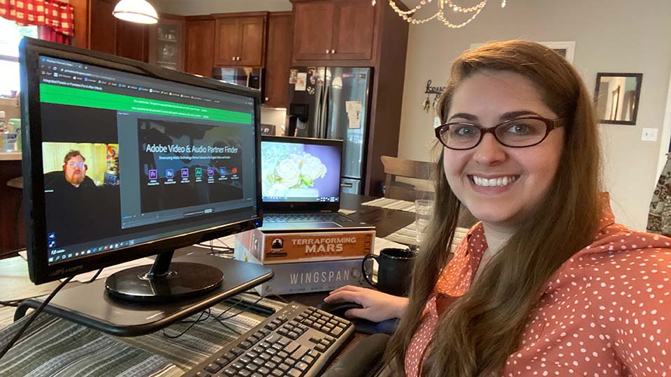 Woman with glasses working from home at dining table with webinar on monitor on riser and laptop on board games